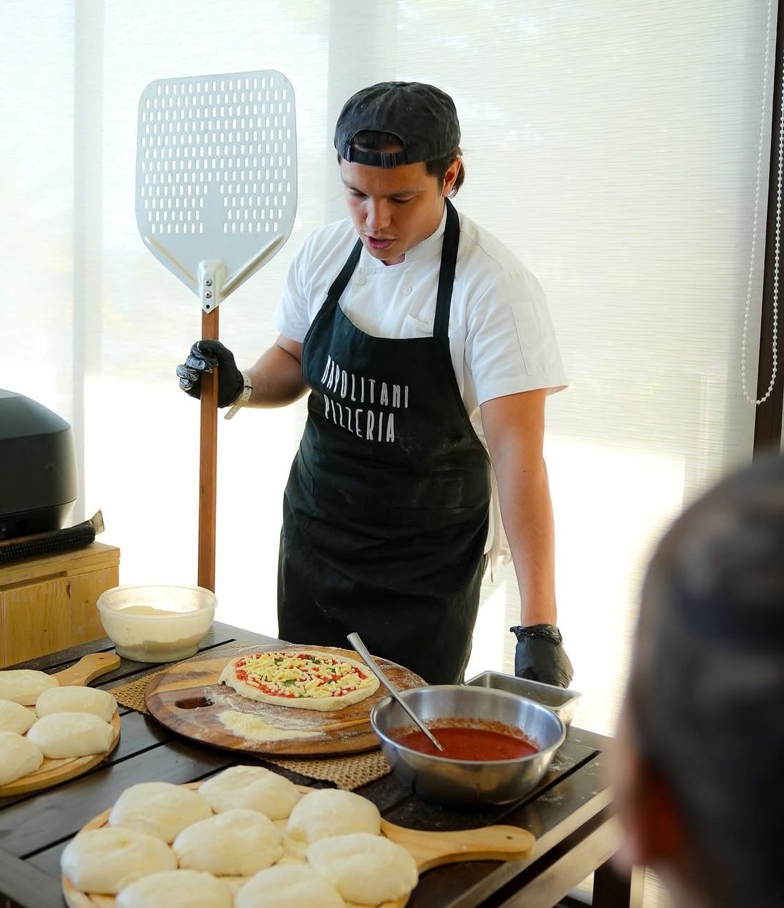 Our pizzaiolo in a Napolitani apron, with dough balls, tomato sauce and a pizza ready for the oven