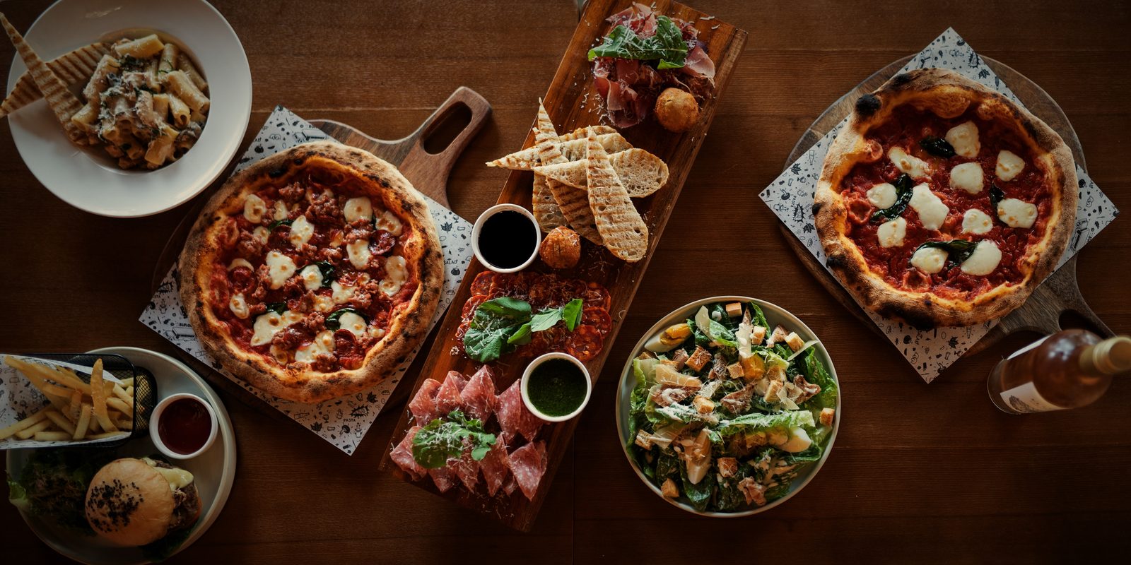 A table spread from above: two pizzas, a charcuterie board with grilled bread, rigatoni and a Caesar salad